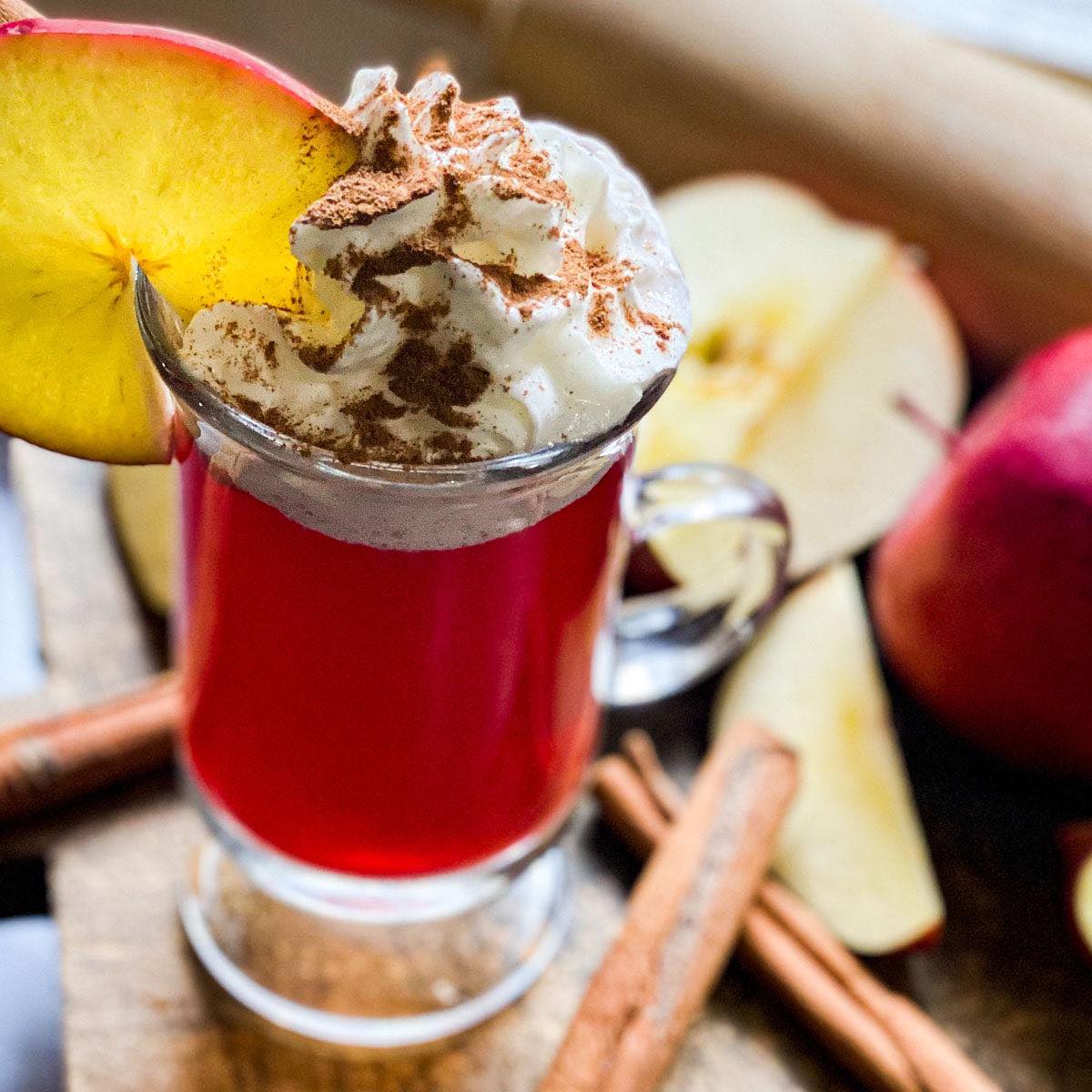 A bright red brewed cup of apple pie tea topped with whipped cream, cinnamon, and apple slice sits on a wooden counter.