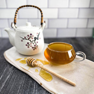 A clear glass tea cup filled with brewed Cough Tea rests on a white ceramic surface with a honey wand and tea pot.