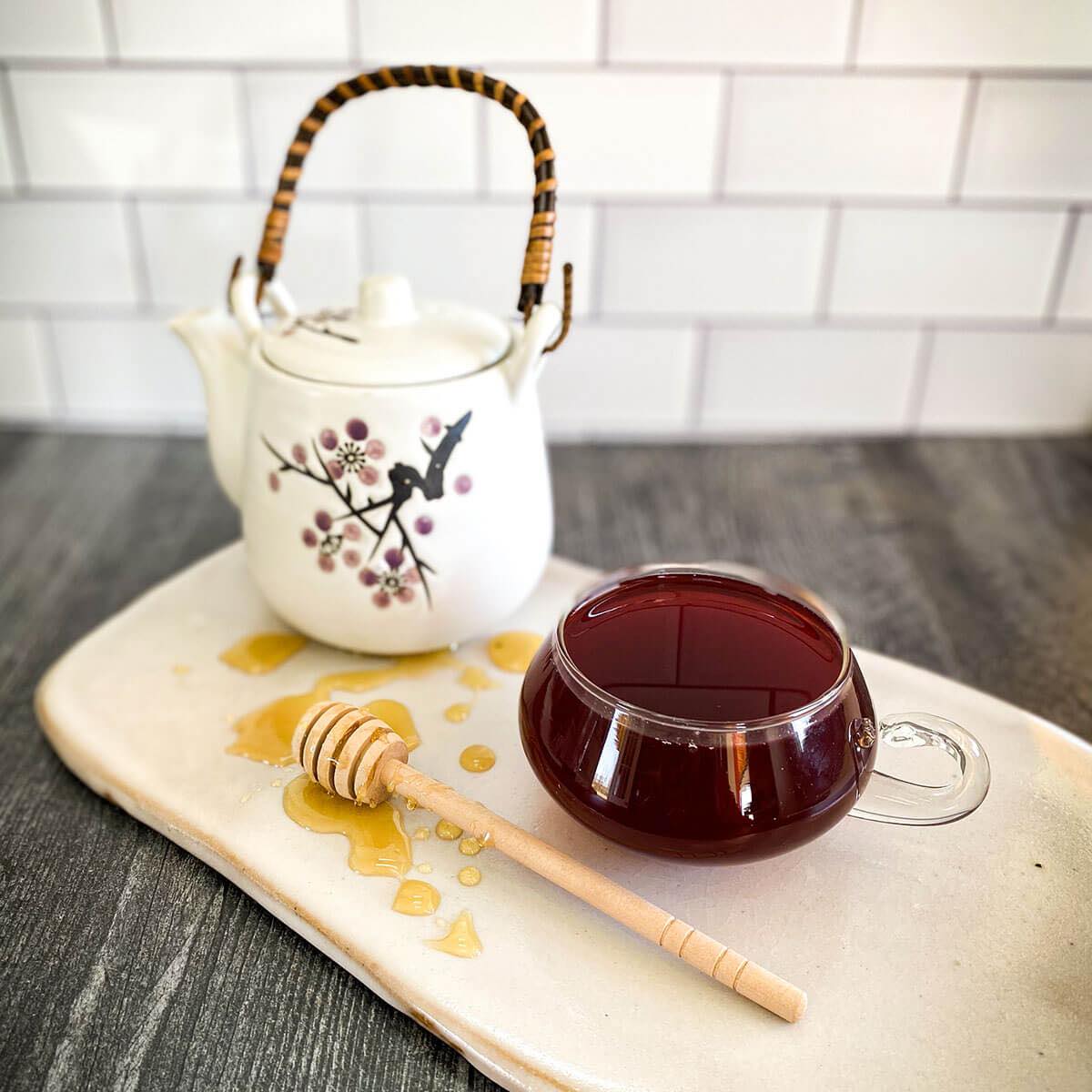 A brewed mug of Herban C tea rests on a white ceramic surface with a tea pot and honey wand.