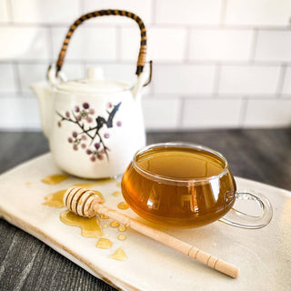 A mug with brewed Rise and Shine Tea rests on a white ceramic surface with a tea pot and honey wand.
