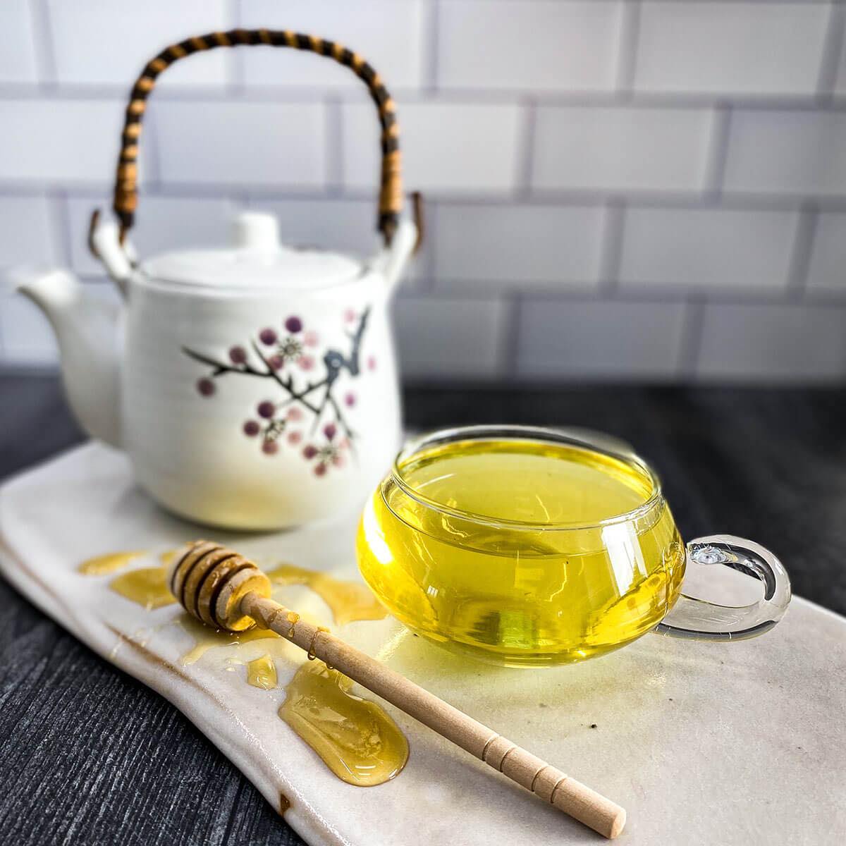 Chamomile tea in a glass mug with a honey wand and tea pot in the background