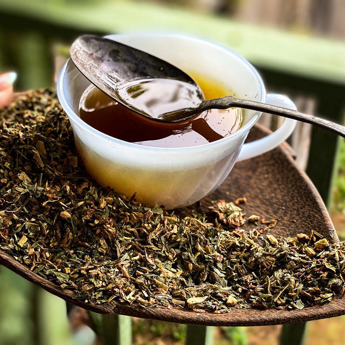 A milk glass tea cup filled with brewed Cough Tea rests on a wooden leaf shaped dish filled with loose leaf tea. A spoon rests on the tea cup.