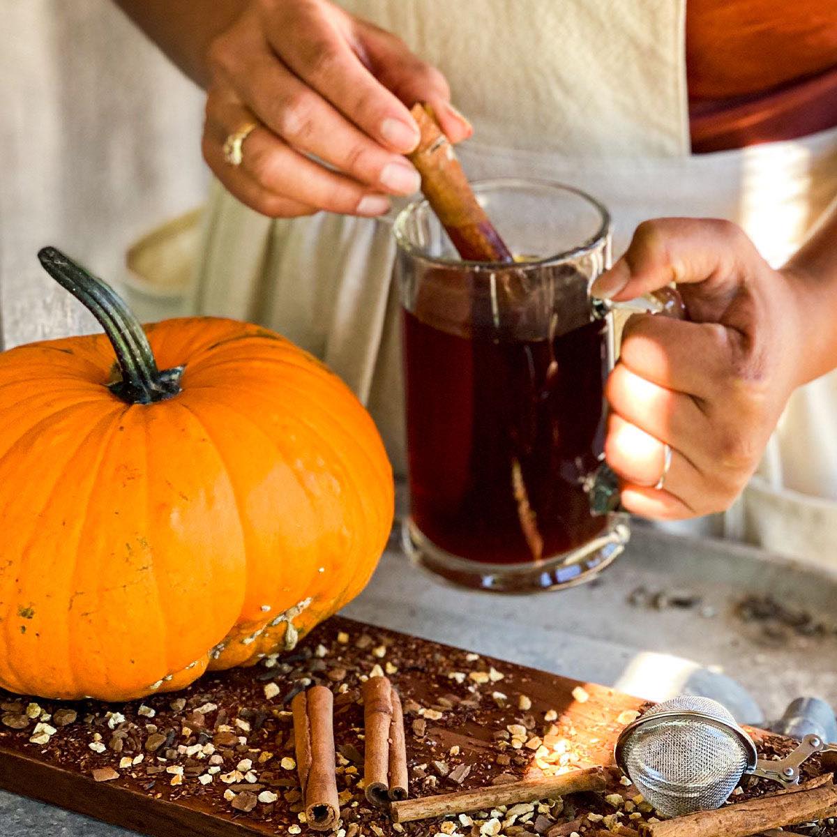 A woman stirs a brewed mug of Creamy Pumpkin Pie tea with a cinnamon stick. A pumpkin and loose leaf tea with a tea strainer are in the foreground.