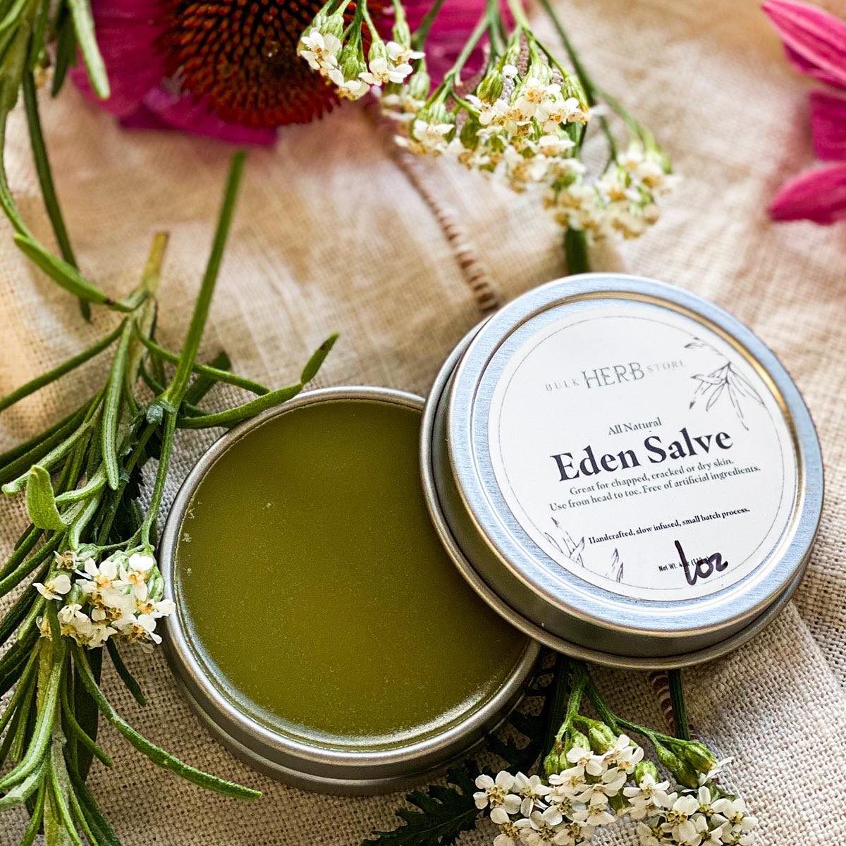 An open tin of Eden Salve rests on a linen cloth with flowers and herbs.