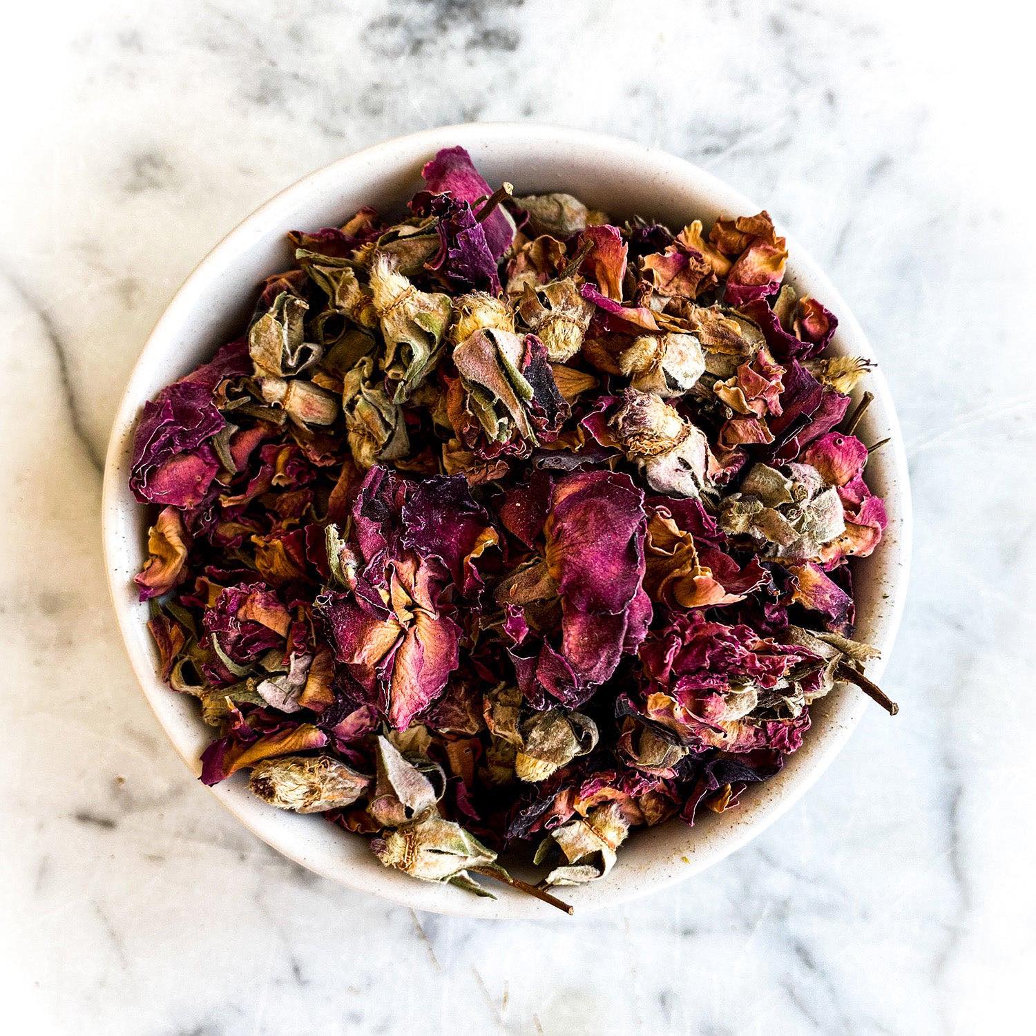 Red rose buds and petals in a white bowl on a white marble background.
