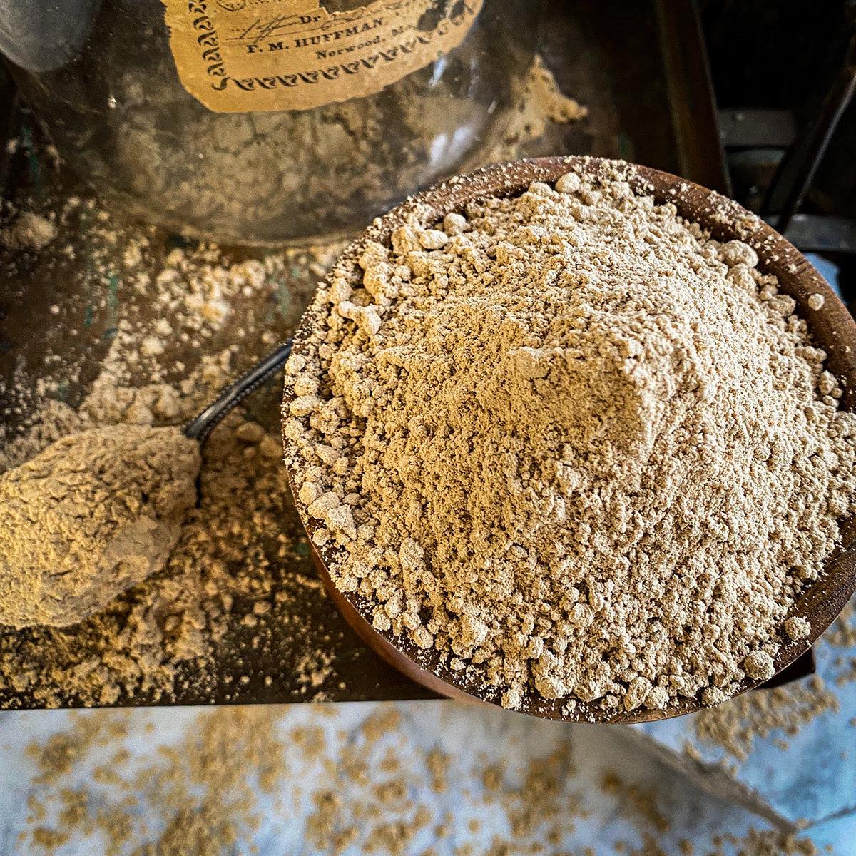 Slippery Elm Bark Powder in a wooden bowl with an old apothecary glass jar in the background and a spoon filled with the powder