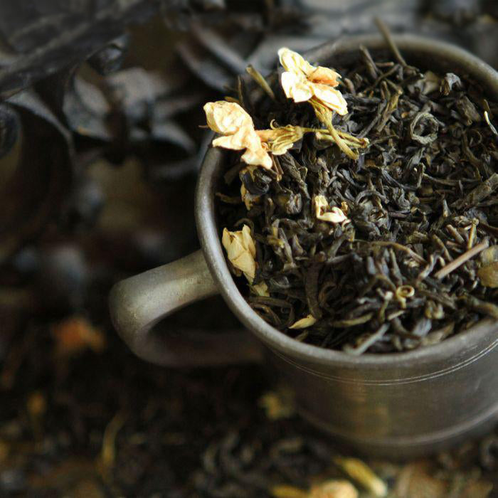 The top view of a dark grey mug overflows with Jasmine Green Tea, with jasmine flowers on top and a blurred background.