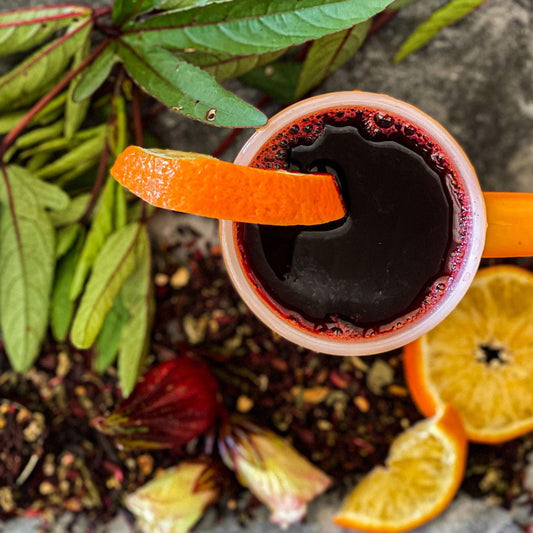 Hibiscus Orange Delight Tea brewed in a mug with a slice of orange on the side surrounded by herbs, flowers and citrus.