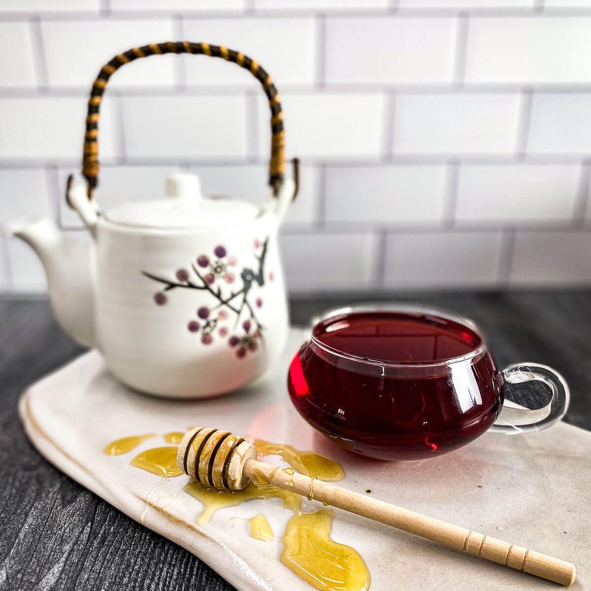 Brewed Hibiscus Orange Delight Tea in a clear glass mug with a white tea pot and honey wand in the background.