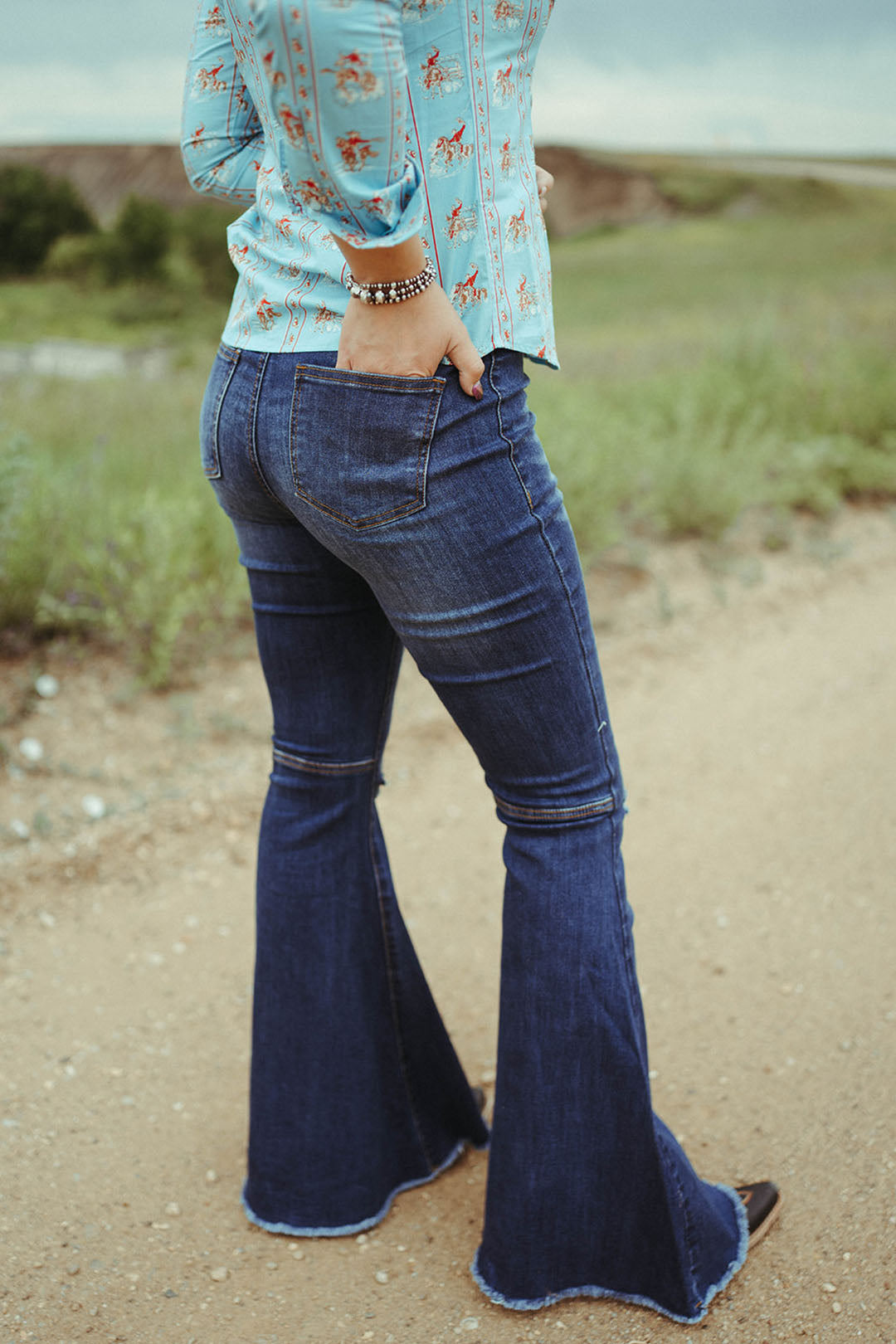 Woman standing on dirt road modeling the back of the Dark Wash Distressed Knee Flares.  2 back pockets. Raw hem. 