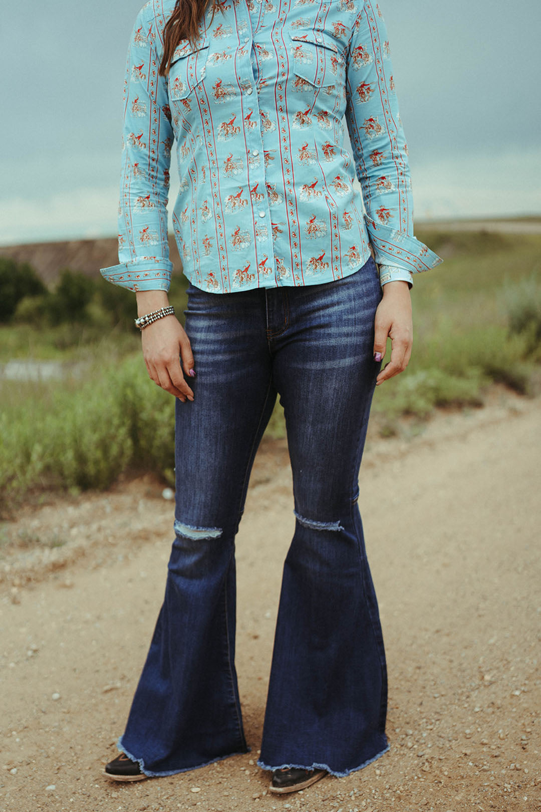 Woman standing on dirt road modeling the Dark Wash Distressed knee flares.  No front pockets. zip fly.  32.5" inseam