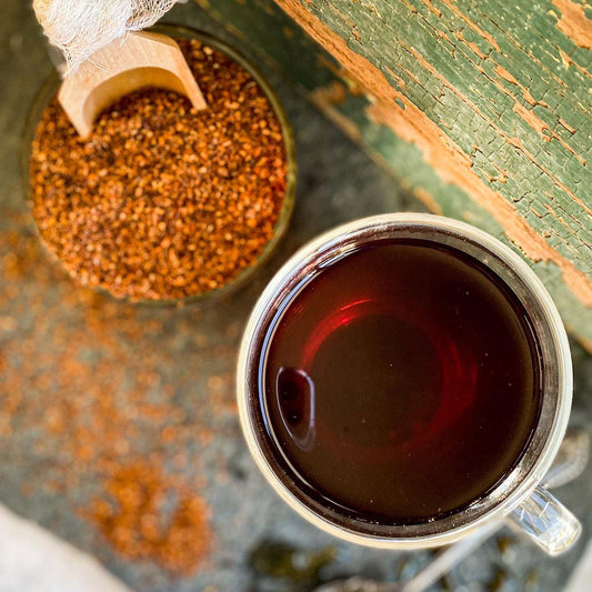 In the background a cup of dried red rooibos with a wooden spoon. In front of the dried tea is a brewed cup of red rooibos.