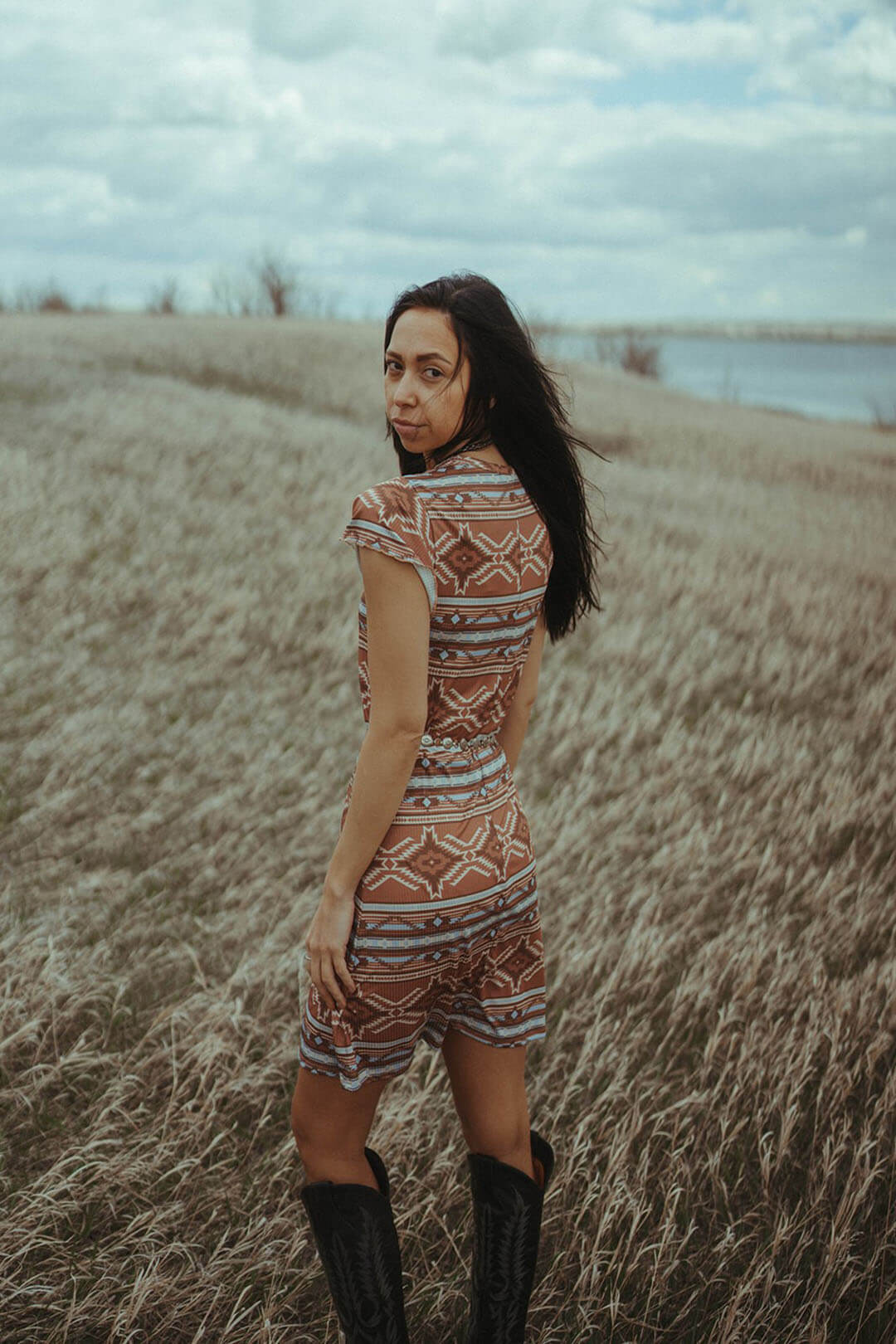 Woman standing in field wearing the Camel Aztec Ribbed Dress.  The dress has colors of browns, tans, and light blue.  