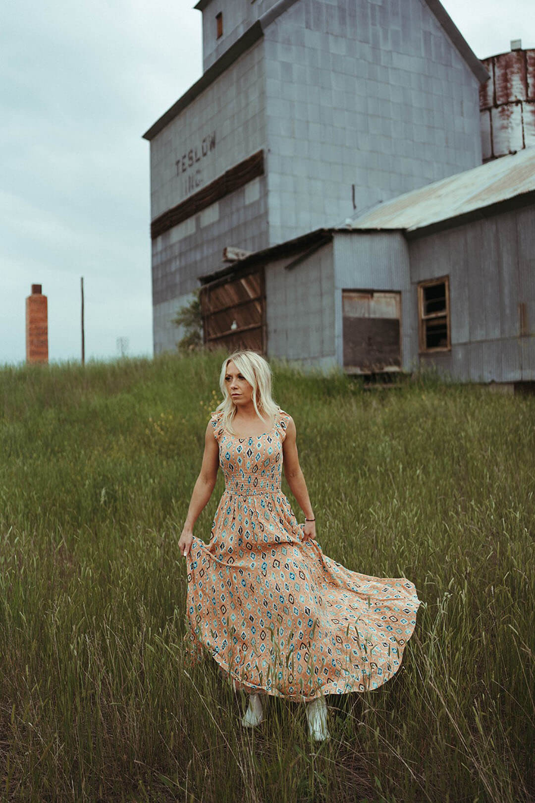 Woman standing in field wearing the Aztec Tiered Maxi Dress by Rock & Roll Denim.  Aztec design throughout.  Orange colored dress. 