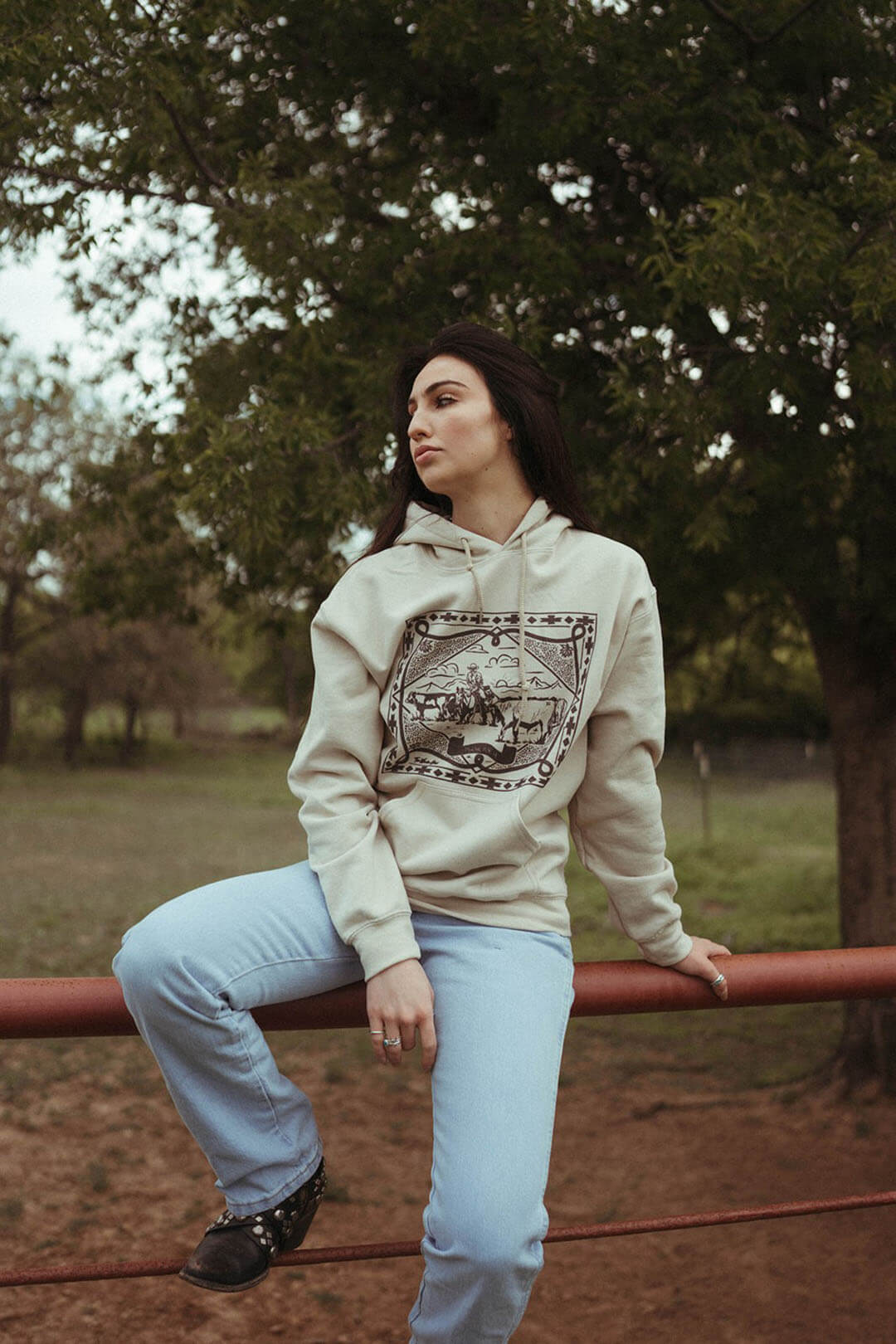 WOman sitting on fence wearing the Psalms 104:14 Sand Hoodie.  Features a western scene on it. 