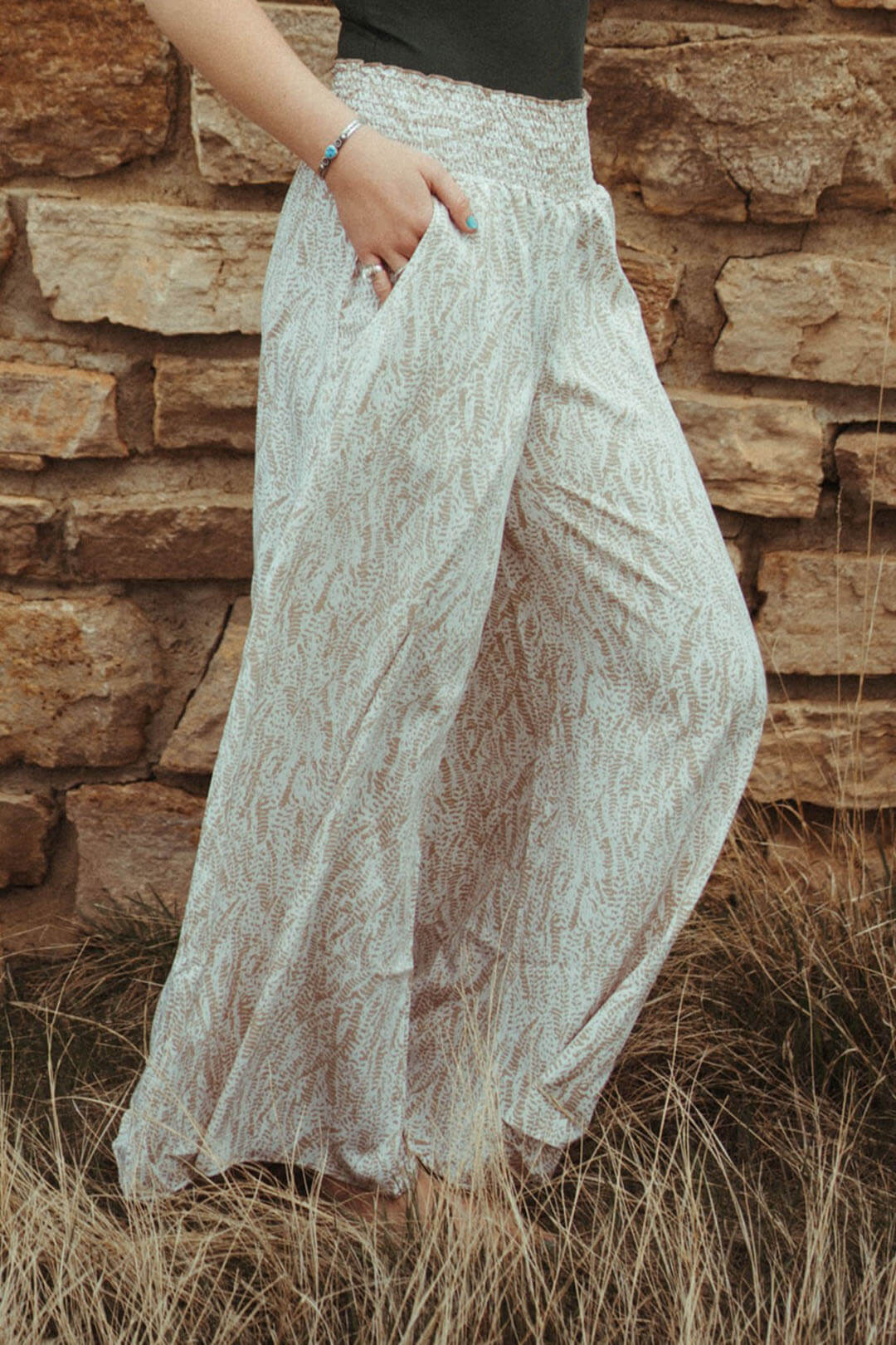 Woman standing next to rock wall modeling the Smocked Waist Maxi Pants.  Flowy and pockets on both sides.  