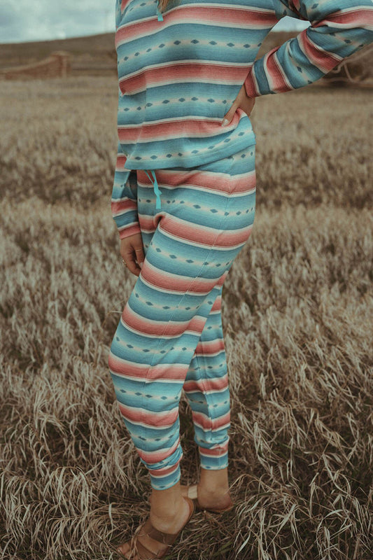 Woman standing in field wearing the Turquoise Serape Joggers by Rock & Roll Denim.  Stripe blue and rose colors.