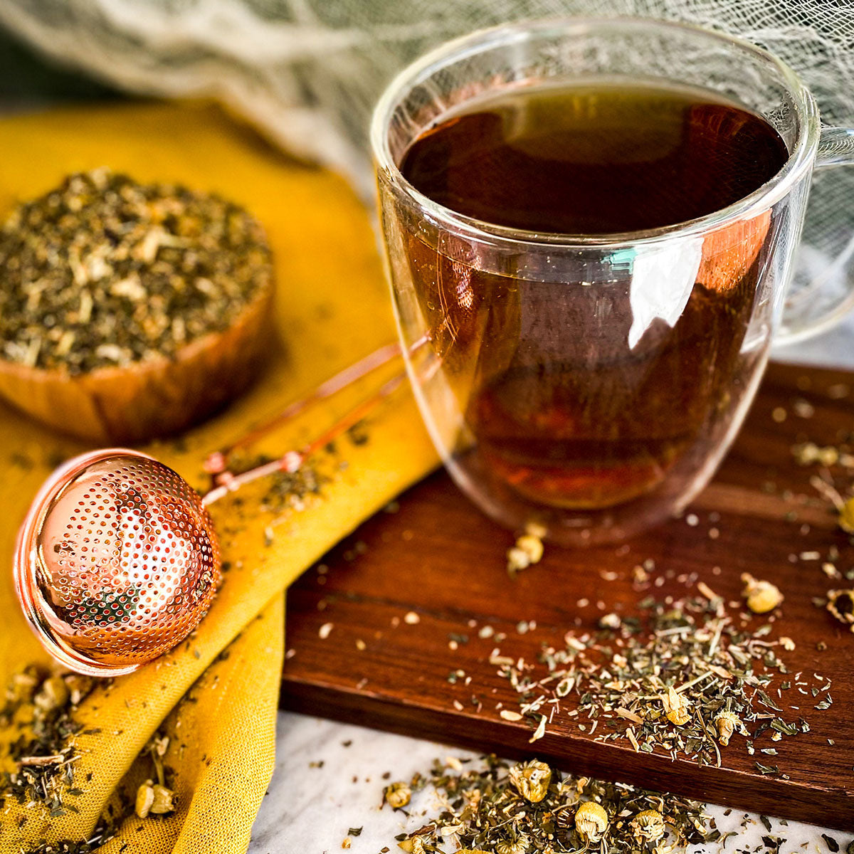 A rose gold tea infuser and brewed cup of cold comfort tea on a wooden surface with loose leaf tea sprinkled on the table.