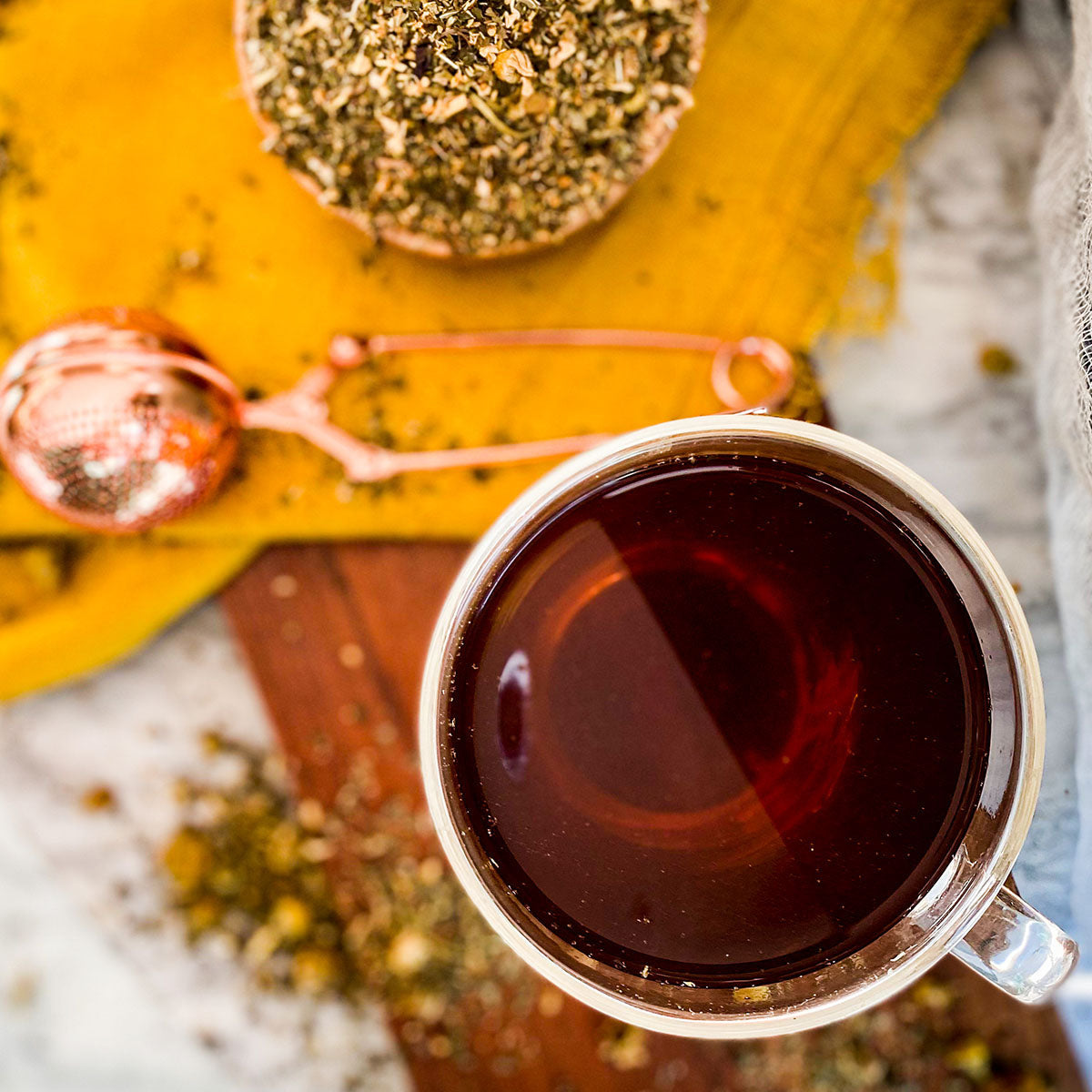 A brewed cup of Cold Comfort tea is surrounded by a yellow napkin and tea ball infuser with loose leaf tea on the table flowing out of a bowl.