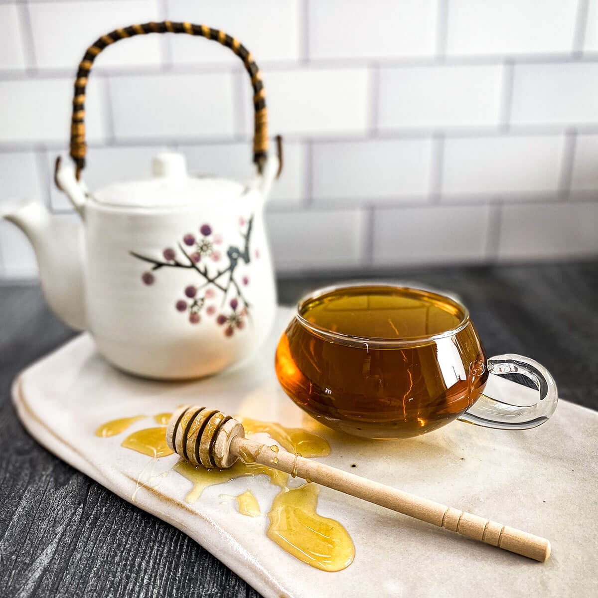 A glass mug of brewed Cold Comfort tea on a counter with a white tea pot and honey wand.