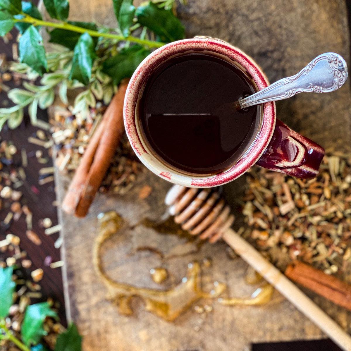 A brewed cup of Christmas Tea in a red mug with silver spoon sits on a wood cutting board with dried herb and greenery in the background.