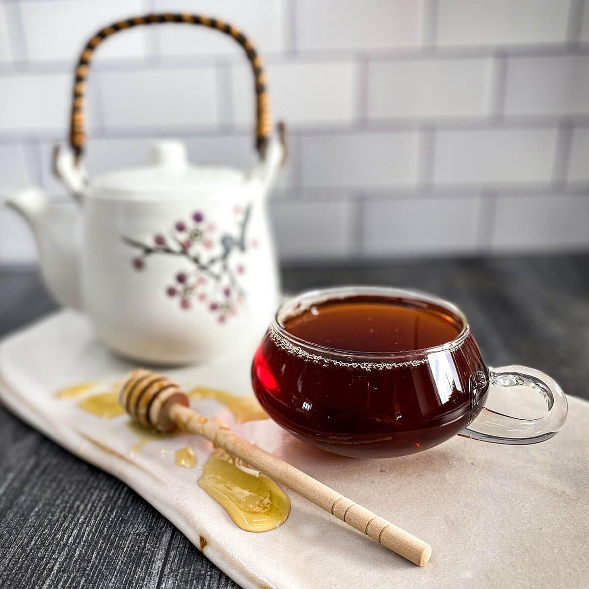Brewed black keemun tea in a clear mug with a white tea pot and honey wand in the background
