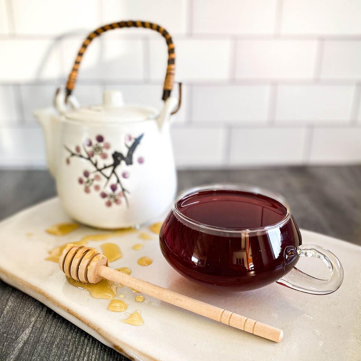 A brewed cup of apple pie tea sits on a counter with a white tea pot and honey wand dripping with honey.