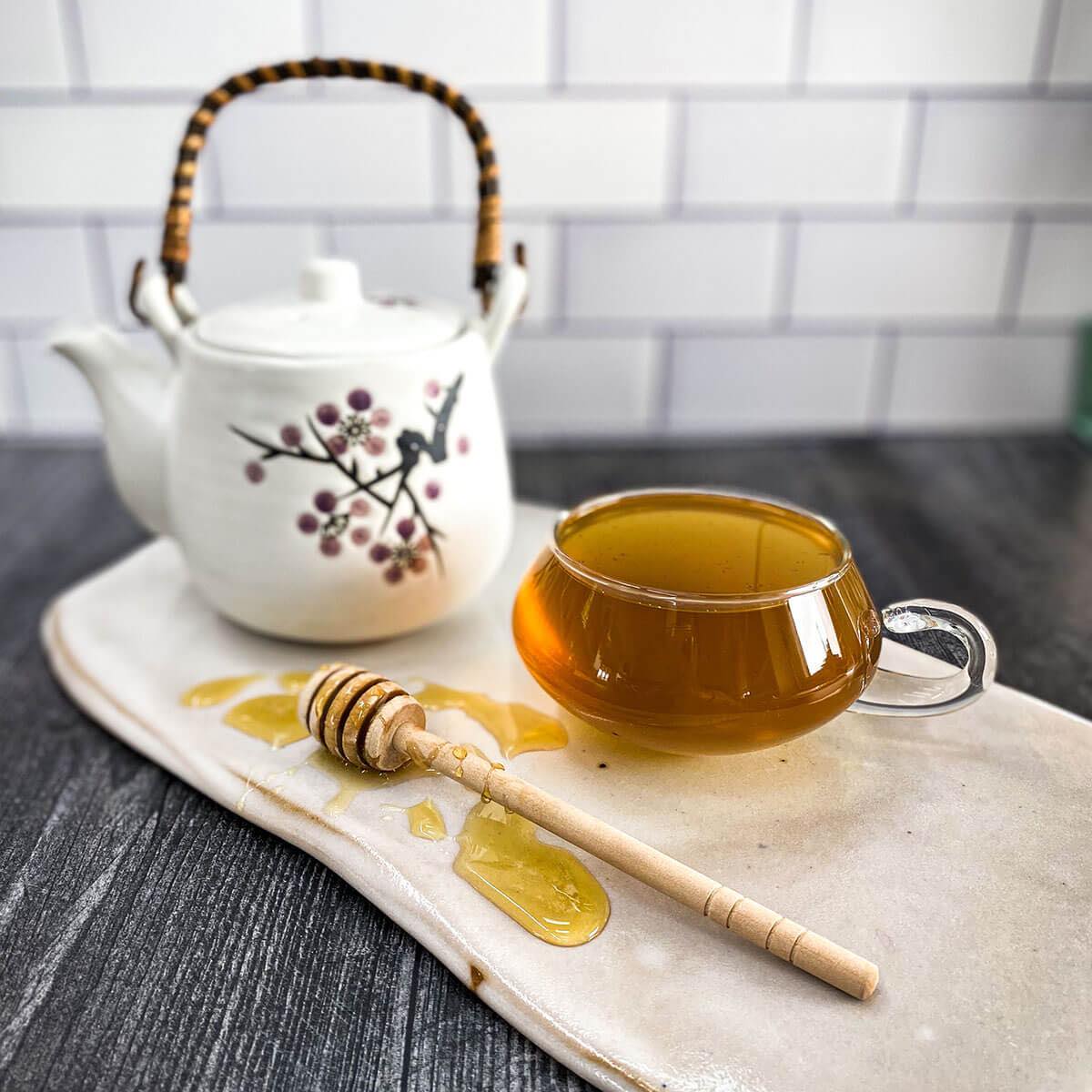 A clear glass tea cup filled with brewed Cough Tea rests on a white ceramic surface with a honey wand and tea pot.