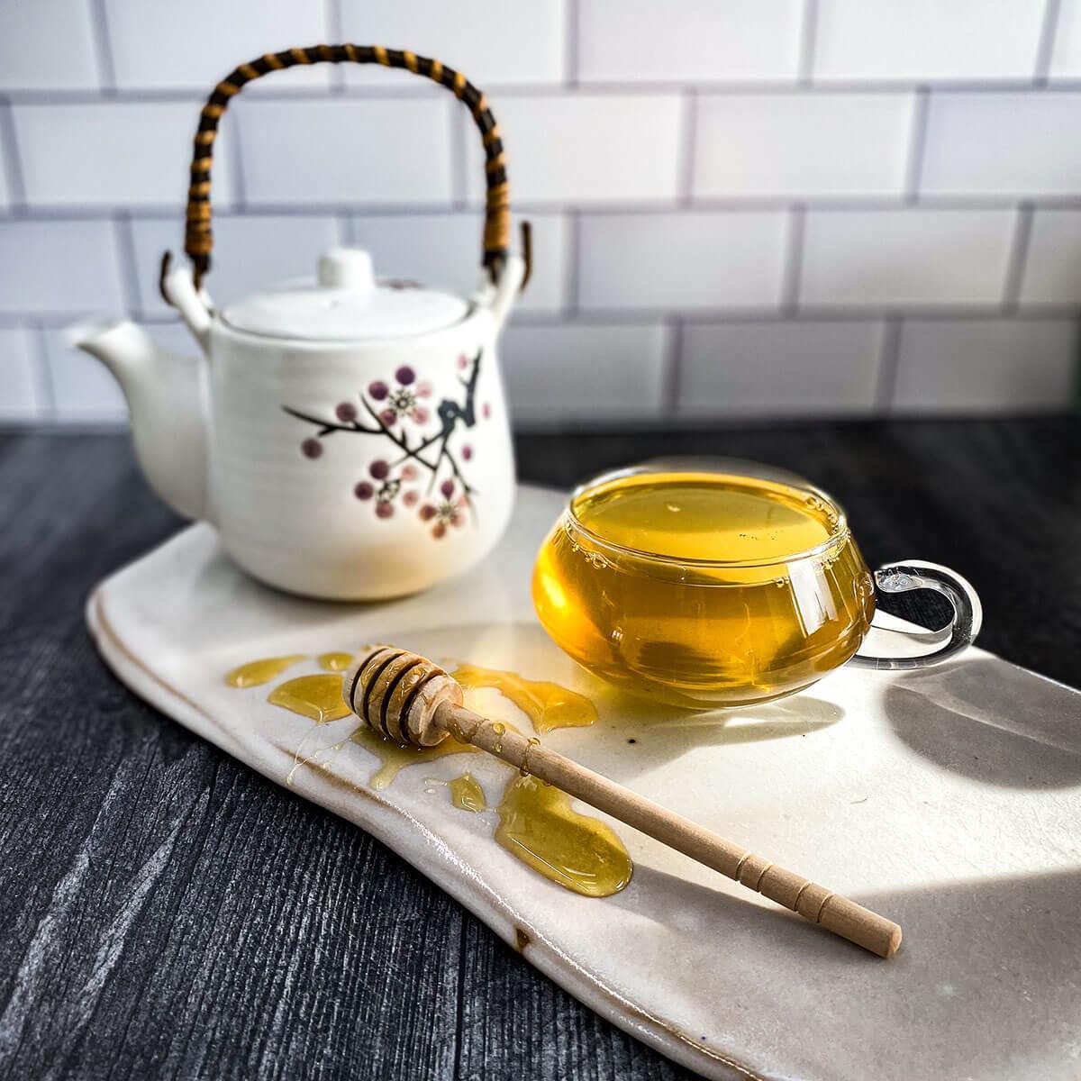 A brewed tea cup of Dr Cinnamon Tea on a white ceramic surface is flanked by a honey wand and white tea pot.