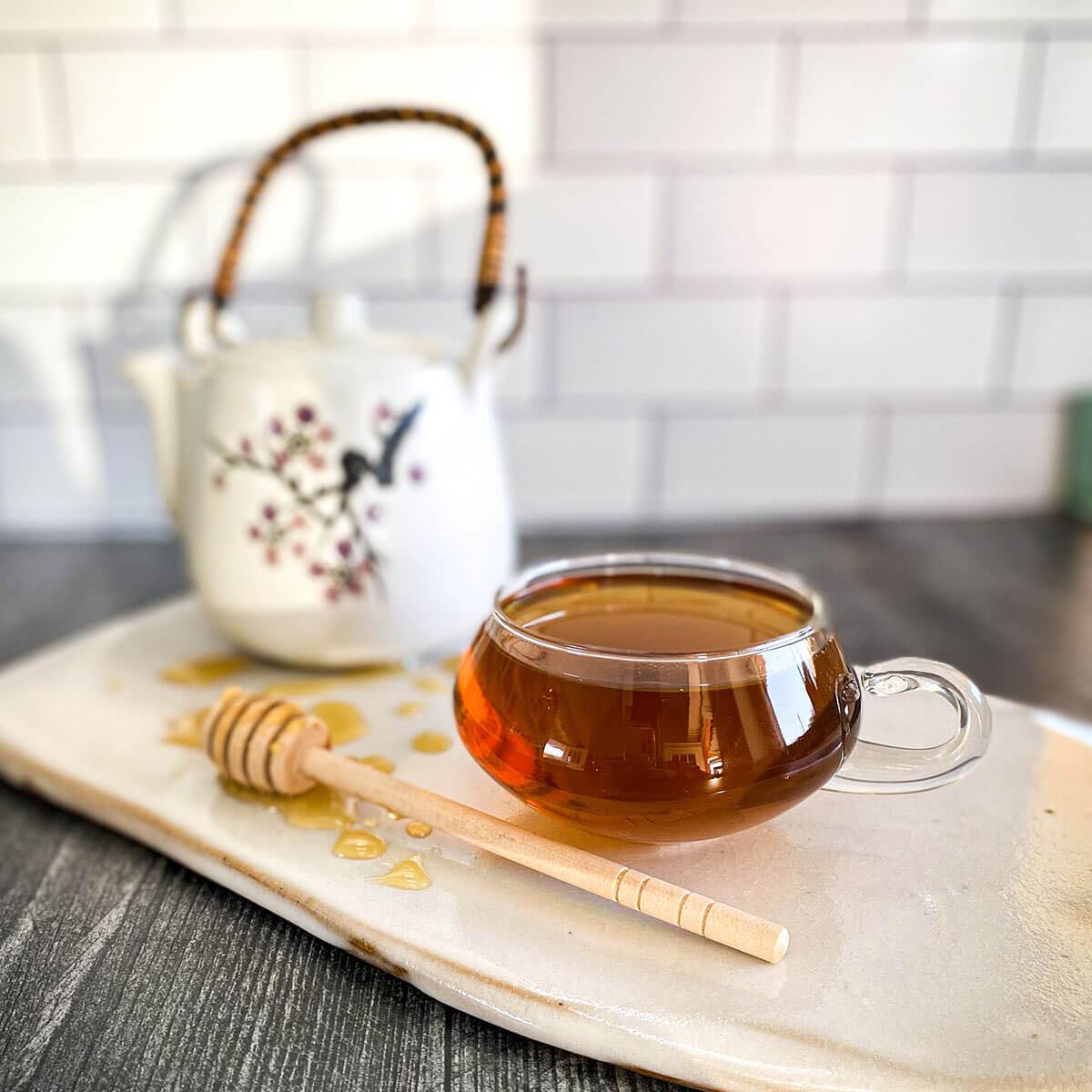 A cup of brewed Mama's Red Raspberry Brew rests on a white ceramic surface with a tea pot and honey wand.
