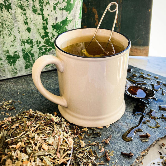 A white mug with brewed Rise and Shine Tea rests on a stone surface with honey and dried herbs.
