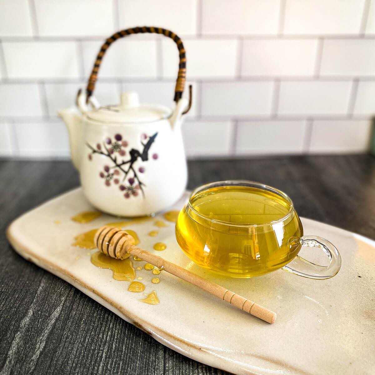 A clear mug of brewed Yerba Mate Tea rests on a ceramic white surface with a white teapot and honey wand.
