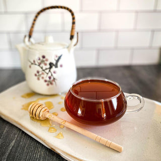 Red Rooibos Spice Chai in glass mug with honey wand in foreground and white tea pot in background.