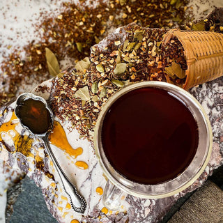 Brewed Red Rooibos Spice Chai in glass mug with honey spoon in foreground and tea strainer with tea spilling out in the background.