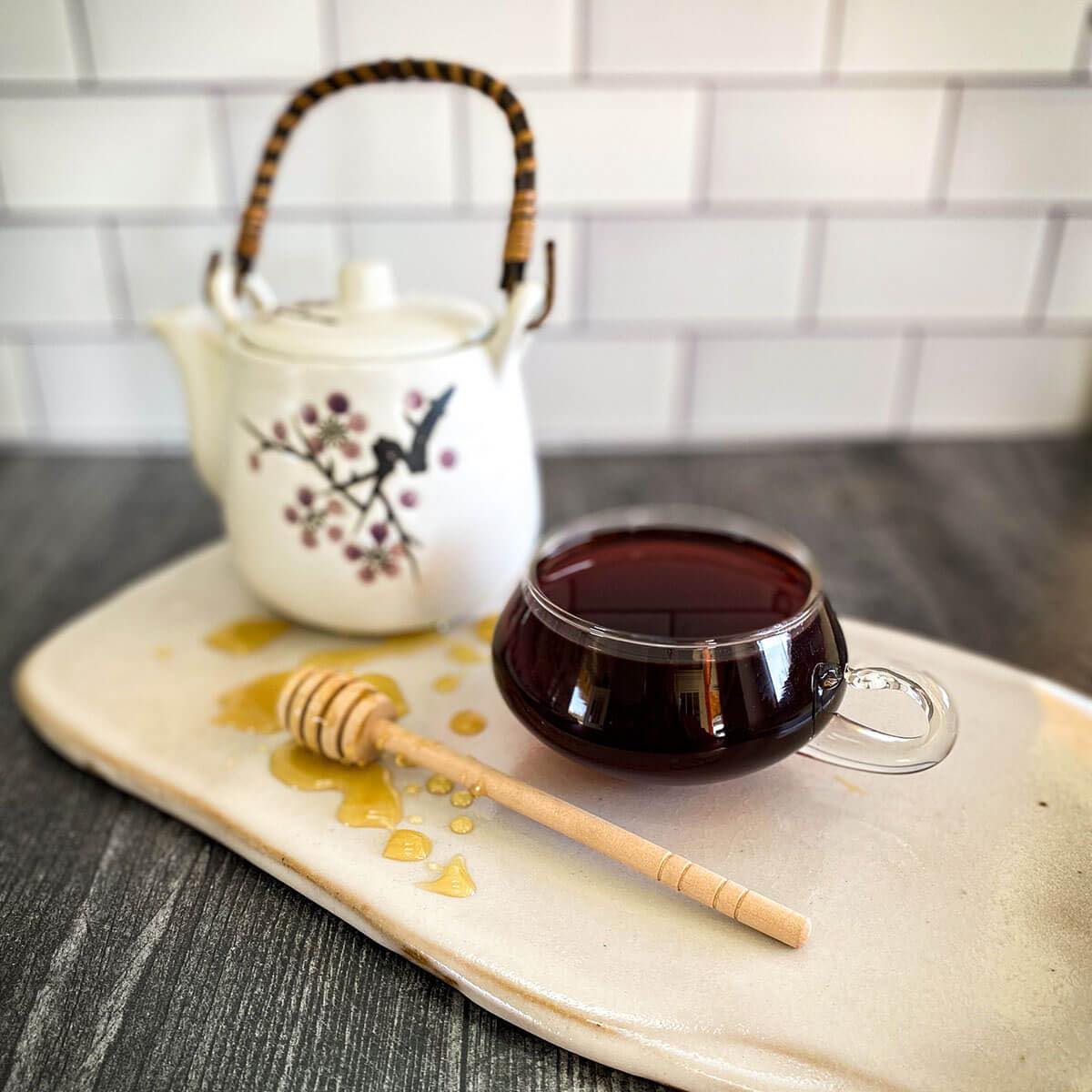 A brewed mug of red rooibos tea is in the foreground with a honey wand dripping with honey and a white tea pot.