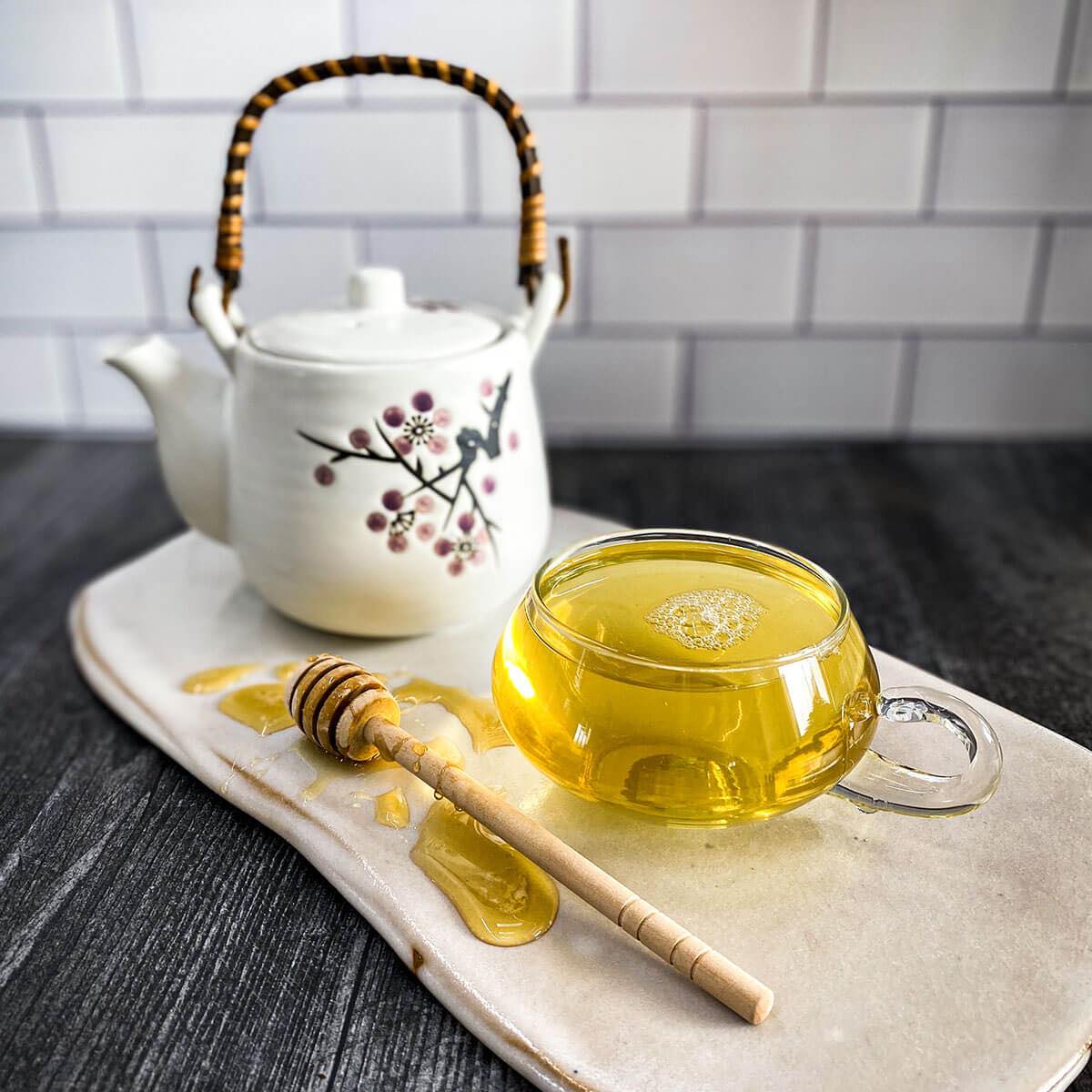 A white tea pot in the background is surrounded by a brewed cup of sencha green tea in a clear mug and a honey wand dripping with honey.