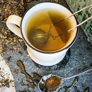 Brewed Rise and Shine Tea fills a white mug with a tea strainer inside. It rests on a stone surface with a spoon of honey in the foreground.