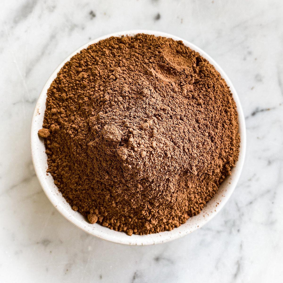 A white bowl filled with cacao powder sits on a white marble table.