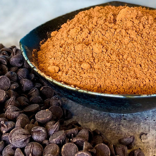 A bowl of cacao powder fills a dish with chocolate chips in the foreground on the table.