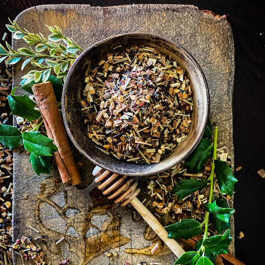 Dried Christmas Tea in a pottery bowl surrounded by cinnamon sticks, honey wand and holly branches on a wood cutting board.