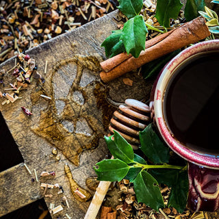 A close up of a red mug of Christmas Tea with holly, a honey wand, and cinnamon sticks on a wooden board.