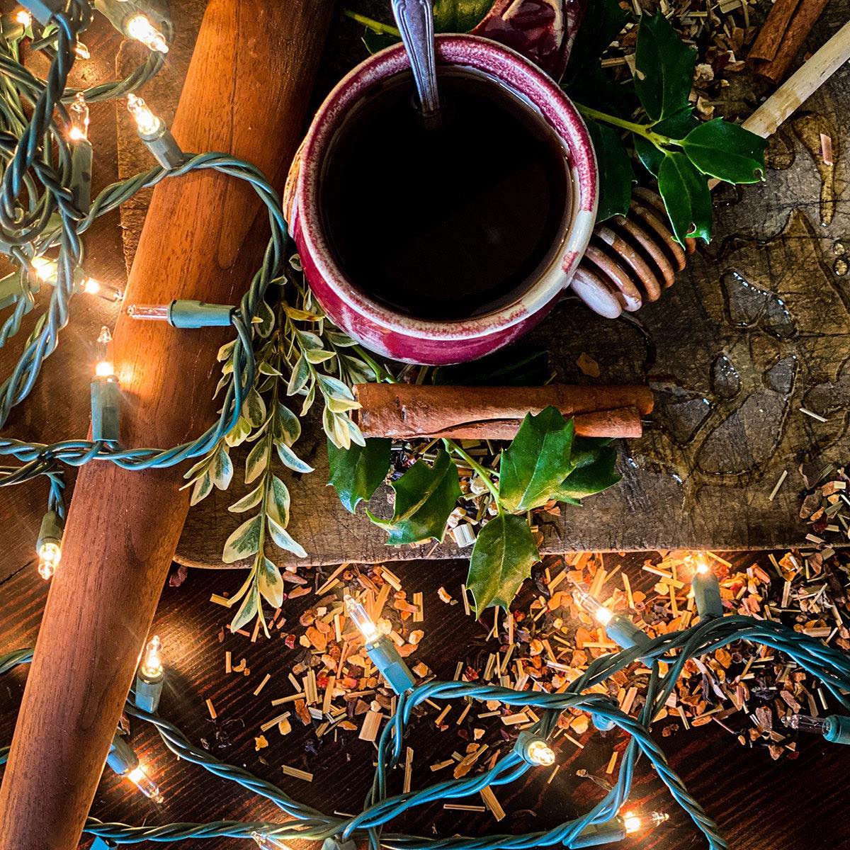 A red mug with brewed Christmas Tea sits on a wooden cutting board surrounded by Christmas lights, greenery and dried herbs.