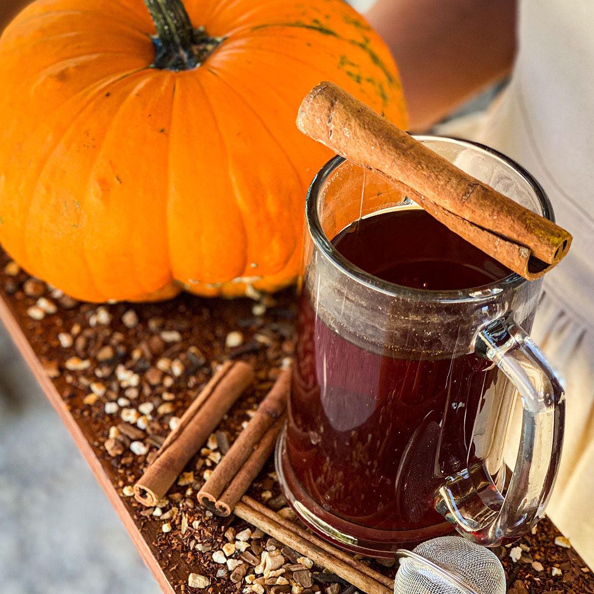 A brewed mug of creamy pumpkin pie tea rests on a wooden surface with cinnamon sticks and sprinkled dried tea. A pumpkin is in the background.