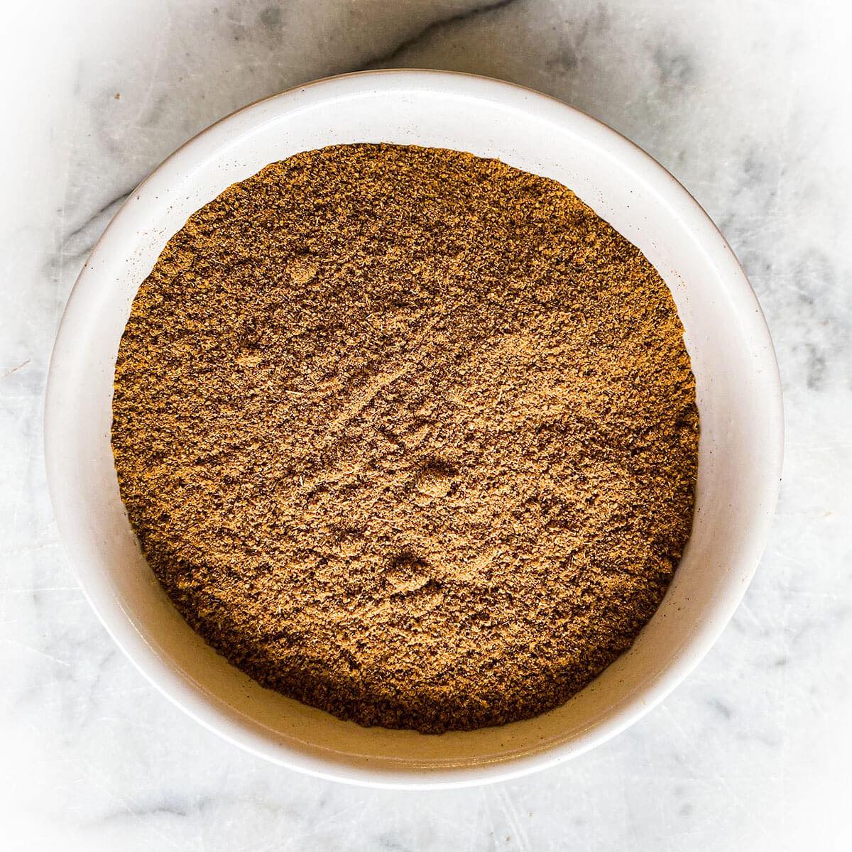 A white bowl with cumin seed powder sits on a white marble table.