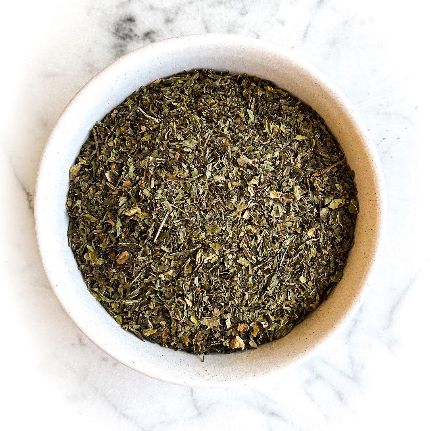 Dried, cut and sifted spearmint leaf in a white bowl on a white marble table.