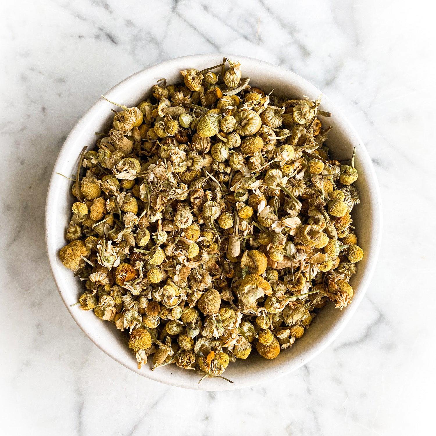 Dried chamomile in a white bowl on a white marble background