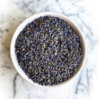 Dried lavender flowers fill a white bowl that rests on a white marble background