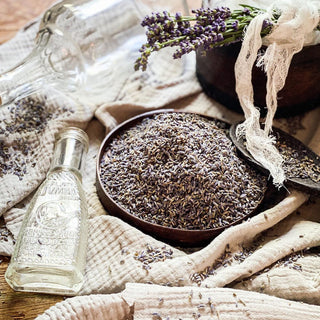 Antique linen as a background with a bowl of dried lavender flowers in the foreground surounded by an antique medicine bottle and wooden spoon.