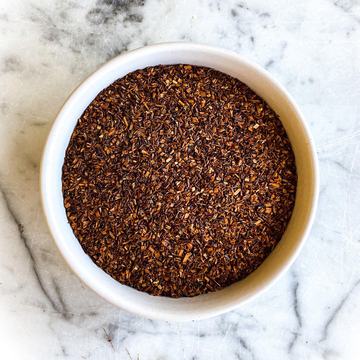 Dried red rooibos tea is featured in a white bowl on a white marble tabletop.