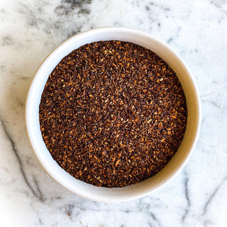 Dried red rooibos tea is featured in a white bowl on a white marble tabletop.